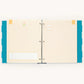 An open three-ring binder planner or notebook with cream-colored lined pages, decorated with teal polka-dot side panels, small yellow tabs, and a small birthday cake illustration on the left page.