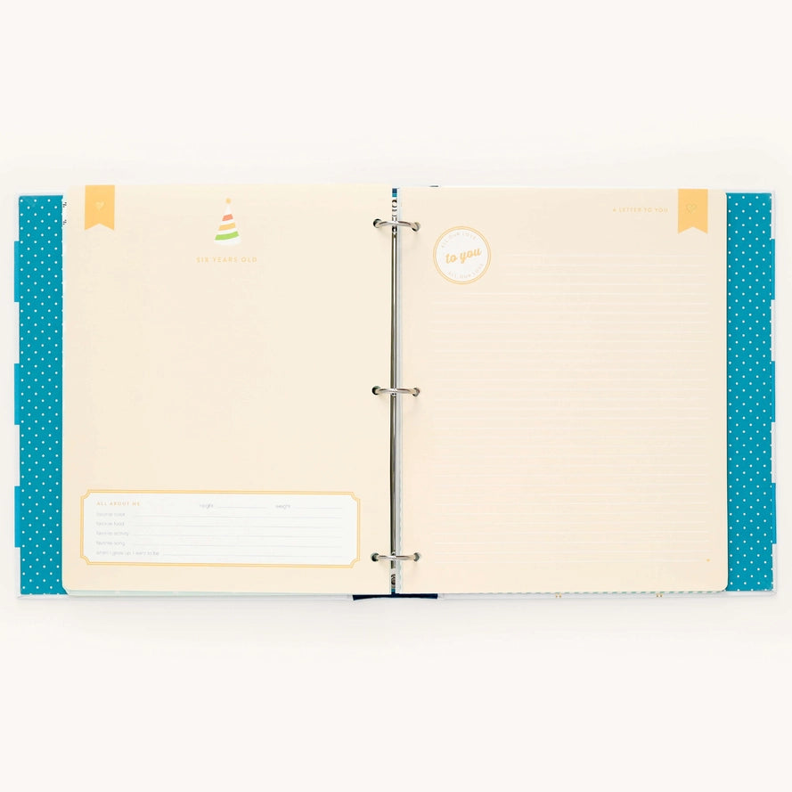 An open three-ring binder planner or notebook with cream-colored lined pages, decorated with teal polka-dot side panels, small yellow tabs, and a small birthday cake illustration on the left page.