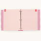A three-ring binder with pink and white patterned pages, featuring diagonal stripes on the main pages, polka dots on the side flaps, and small red heart-shaped tabs on the top corners.