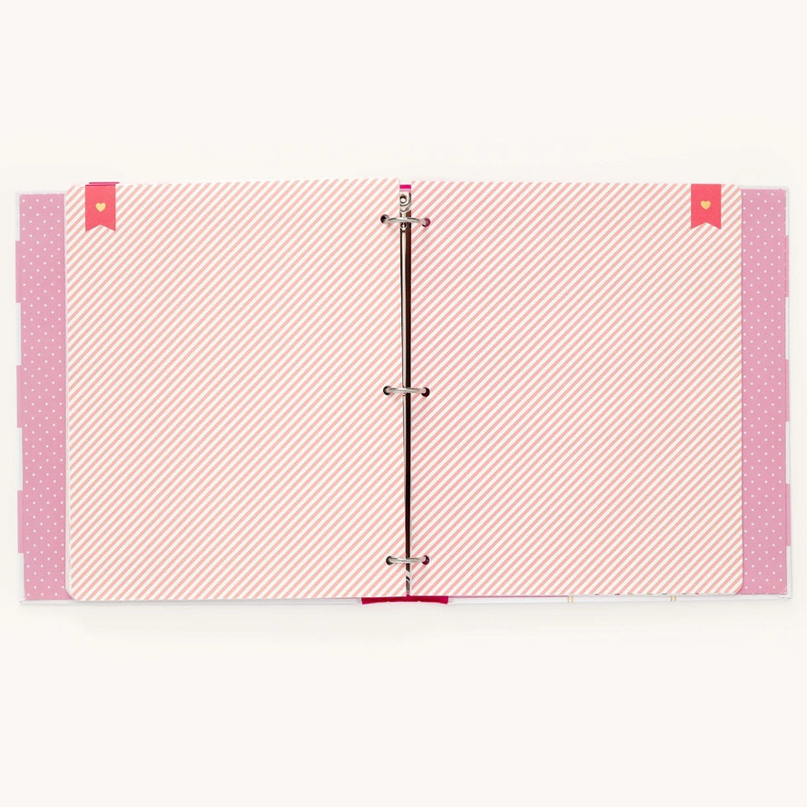 A three-ring binder with pink and white patterned pages, featuring diagonal stripes on the main pages, polka dots on the side flaps, and small red heart-shaped tabs on the top corners.