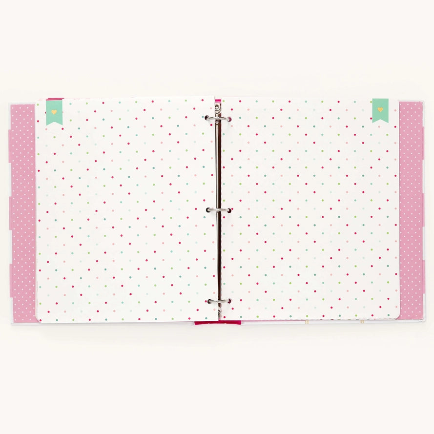 An open three-ring binder notebook with white pages decorated with small pink and green polka dots, pink patterned side tabs, and mint green bookmark tabs on a light cream background.