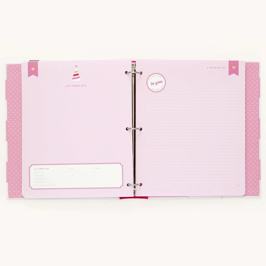 An open pink ring-bound journal or planner binder with lined pages, decorative pink polka-dot side tabs, and small printed birthday-themed illustrations on a plain light background.