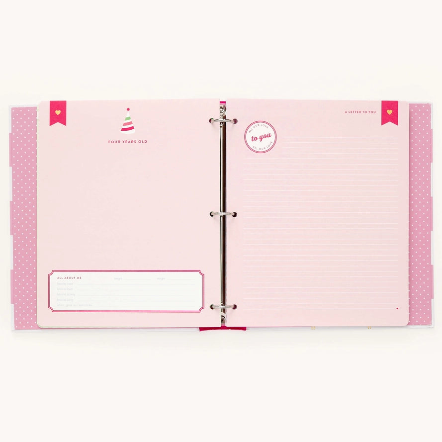 An open pink ring binder planner or keepsake album with lined pages, decorative pink side tabs, and printed heart and circle labels on a light neutral background.