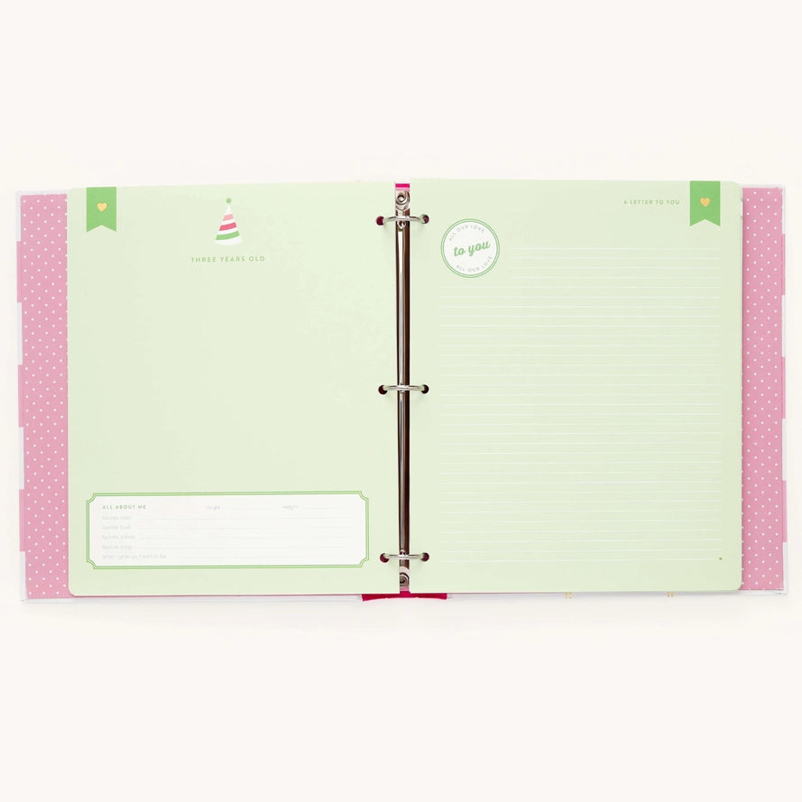 An open three-ring binder planner or agenda with pale green lined pages, pink polka-dot side tabs, and small decorative printed details, photographed on a plain light background.