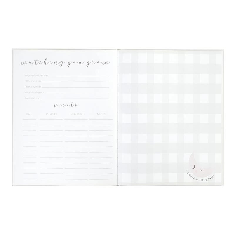 A white baby milestone tracker and photo album shown open, featuring lined spaces for writing and a light gray gingham pattern on the cover, with a small smiling crescent moon illustration in the bottom right corner.